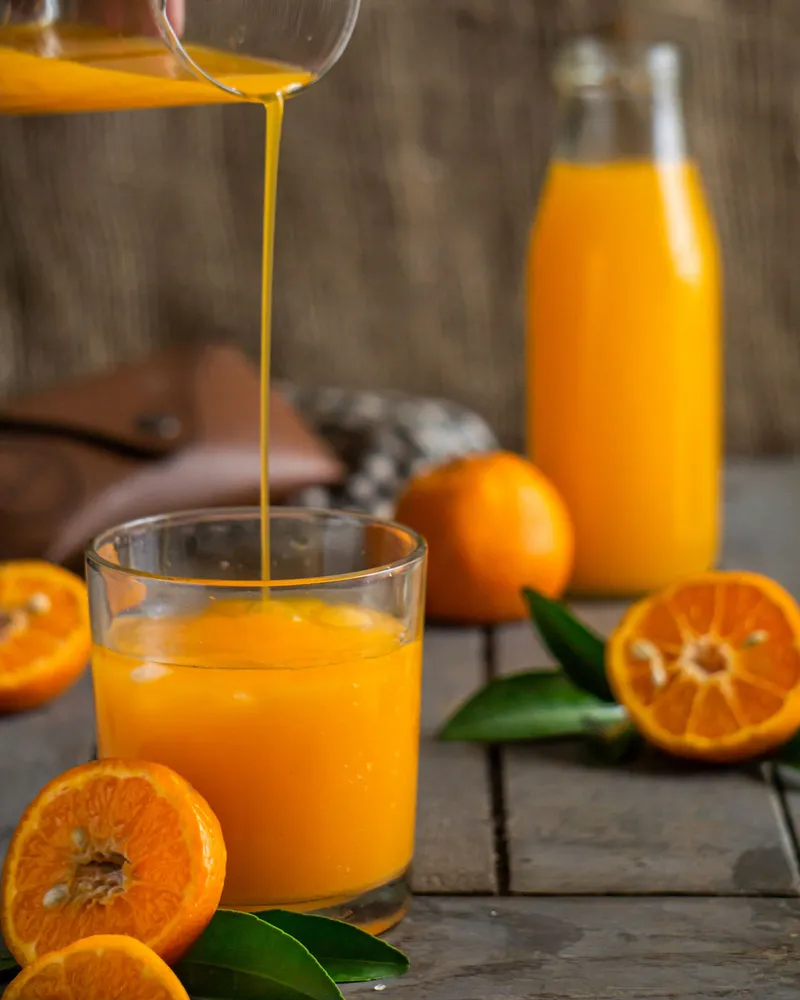 Zumo de naranja recién exprimido cayendo en un vaso rodeado de mandarinas partidas