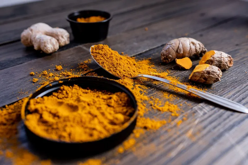 Cucharada de cúrcuma molida junto a raíces frescas de cúrcuma y jengibre sobre madera